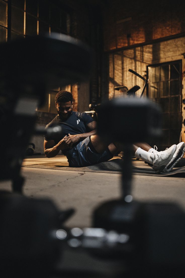 Warm lit gym photo of a man sitting on a mat between dumbbells, catching his breath between sets